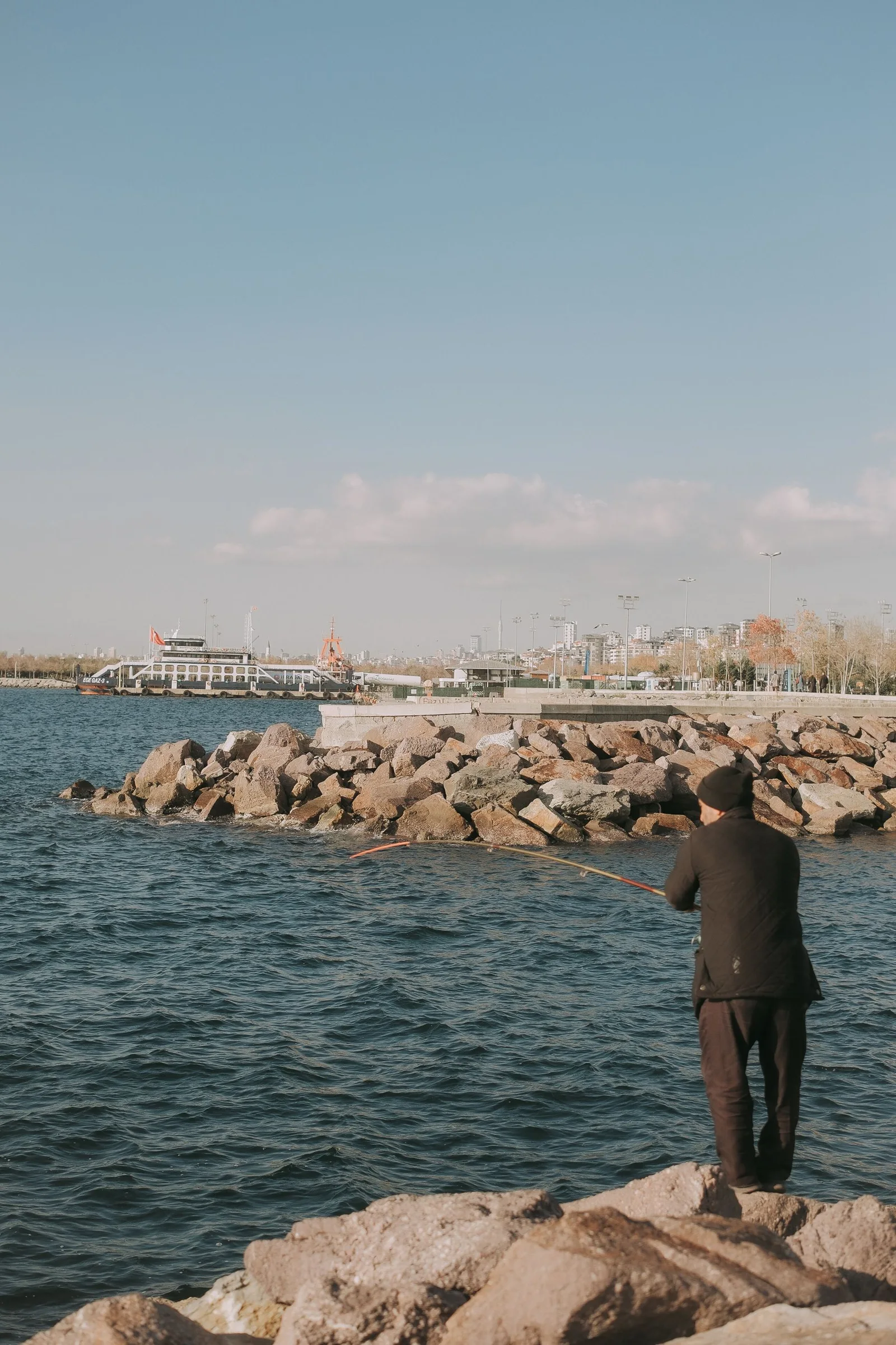 An angler on a rocky waterfront with a city beyond
